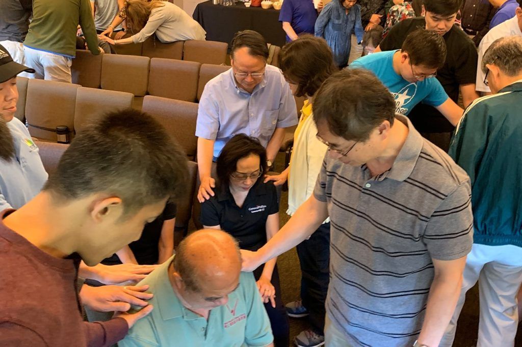 People stand in a circle with hands placed on others’ shoulders during a prayer session inside a meeting room.