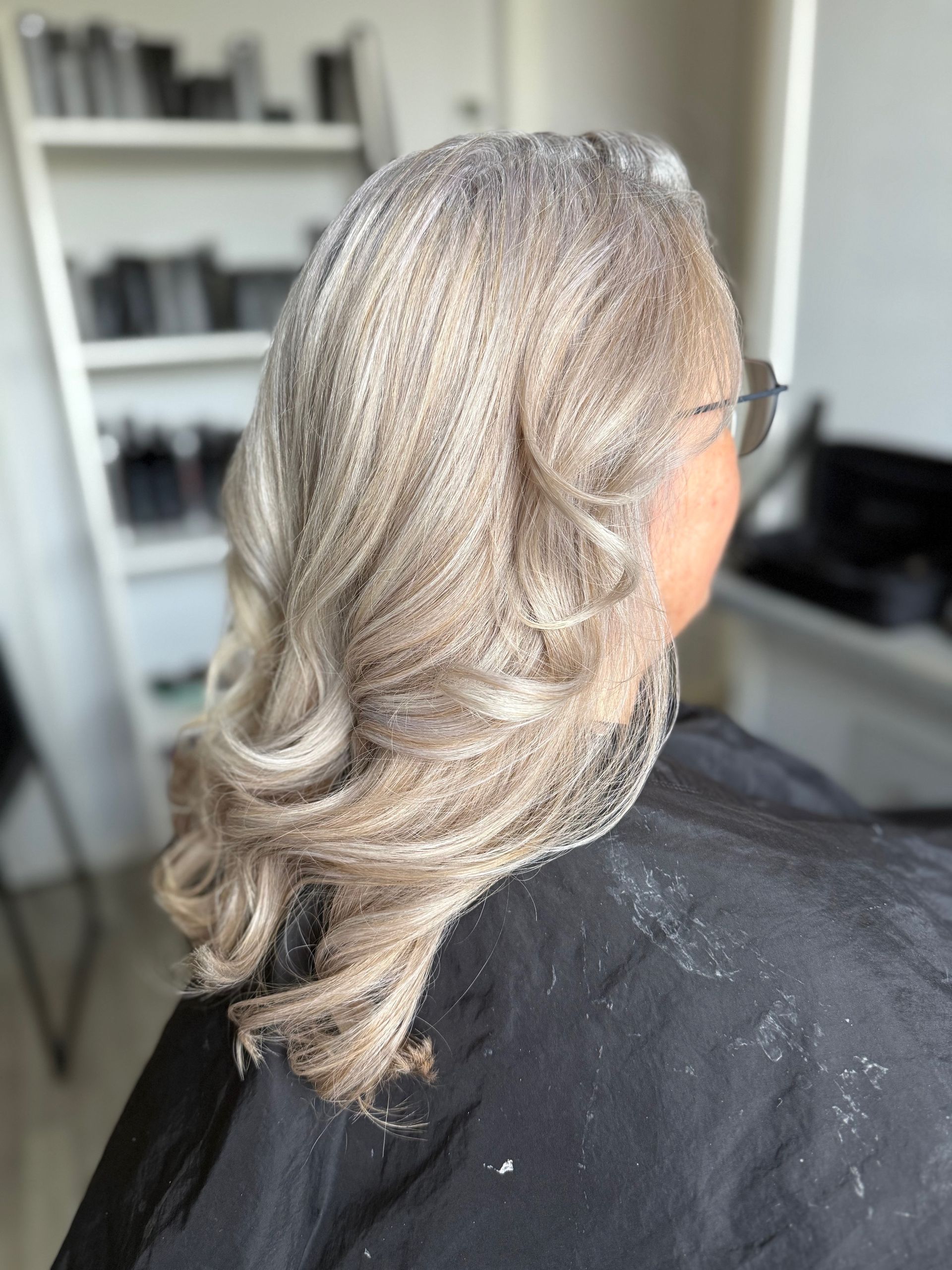 Vrouw met golvend, zilvergrijsblond haar; achterkant. Het haar is gestyled en gedrapeerd over een zwarte cape.