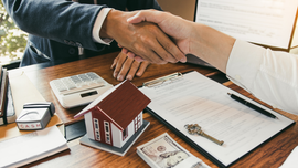 Two people shaking hands over documents, a house model, and cash on a desk, in an office setting.