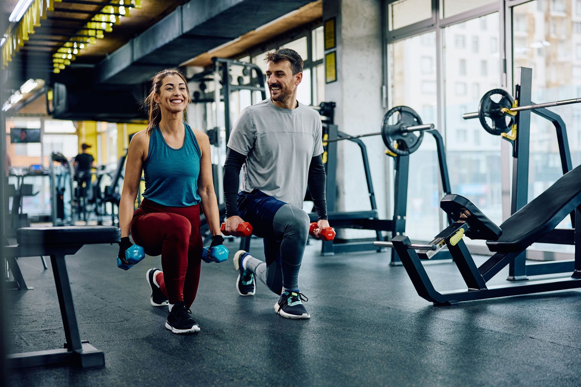 A smiling person in a blue tank top and another in a grey t-shirt perform lunges with dumbbells in a modern gym.