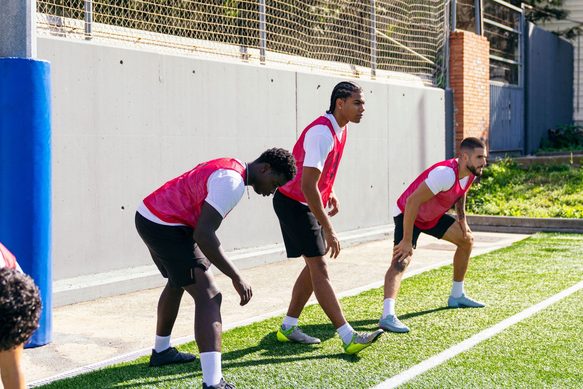 Three athletes in white shirts and pink bibs perform warm-up stretches on an outdoor turf field.