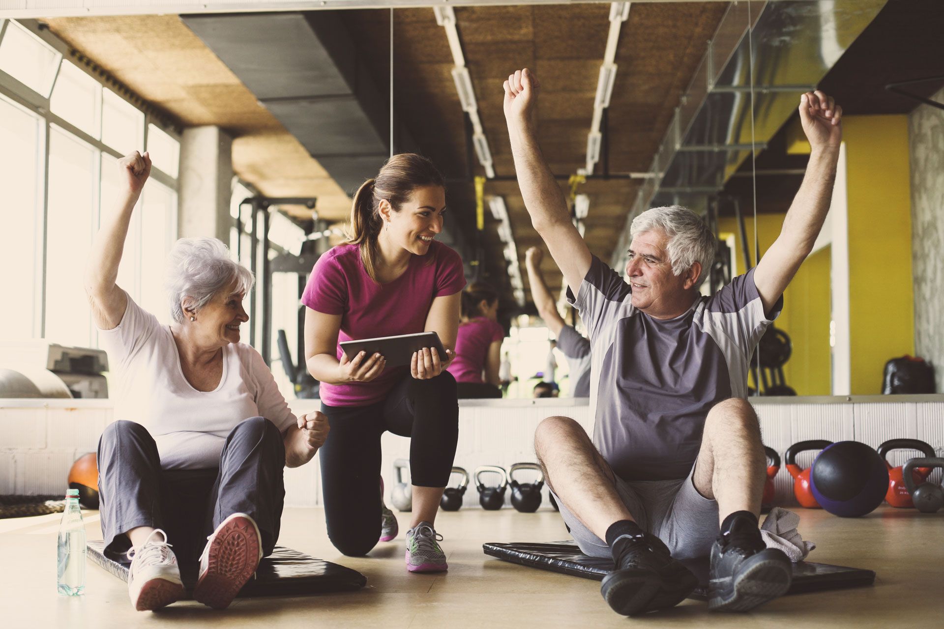 A fitness instructor smiling and holding a tablet while two people sit on gym mats with arms raised in victory.