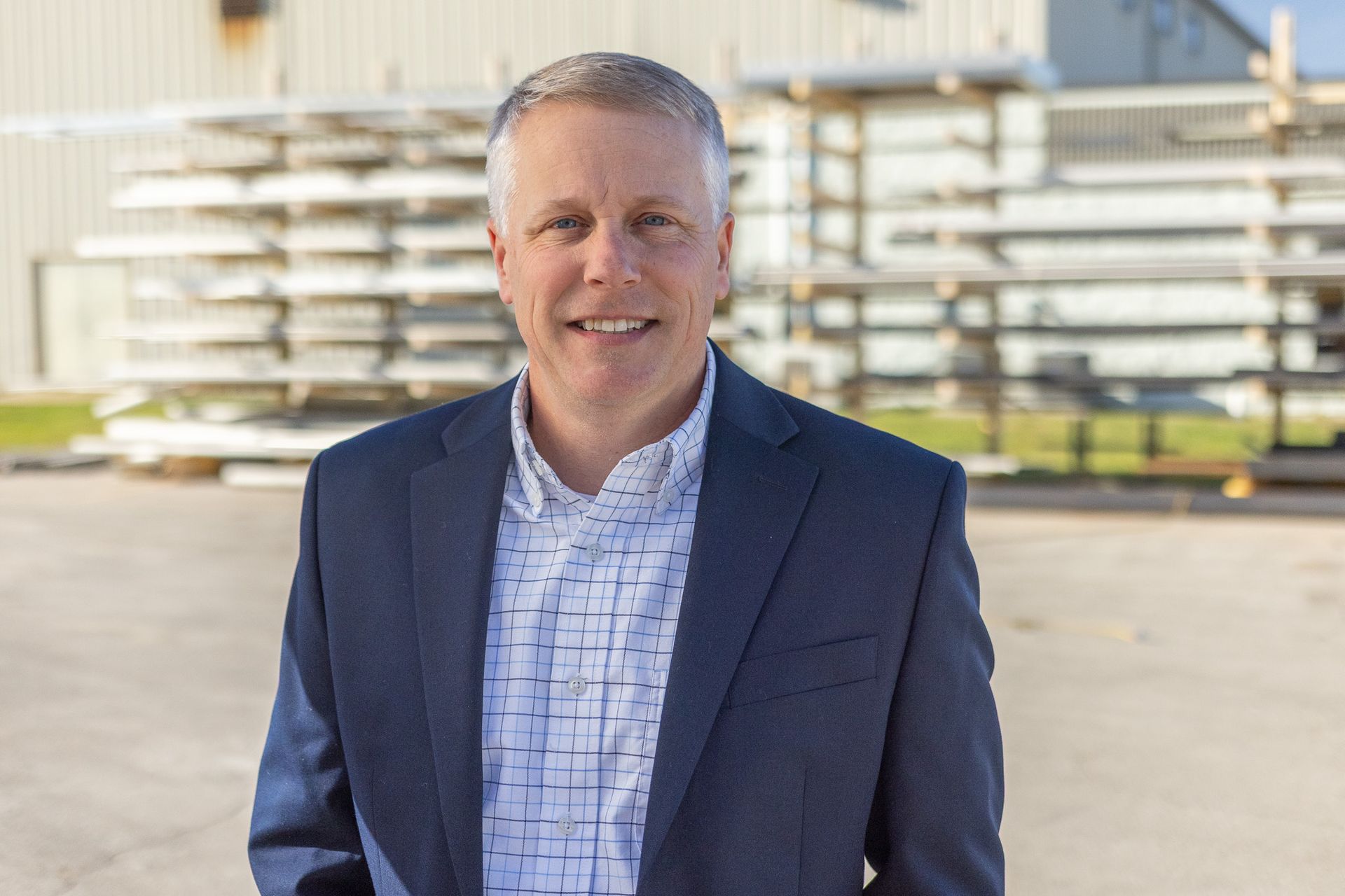 Man with short gray hair smiles, wearing a navy blazer over a white shirt, black background.