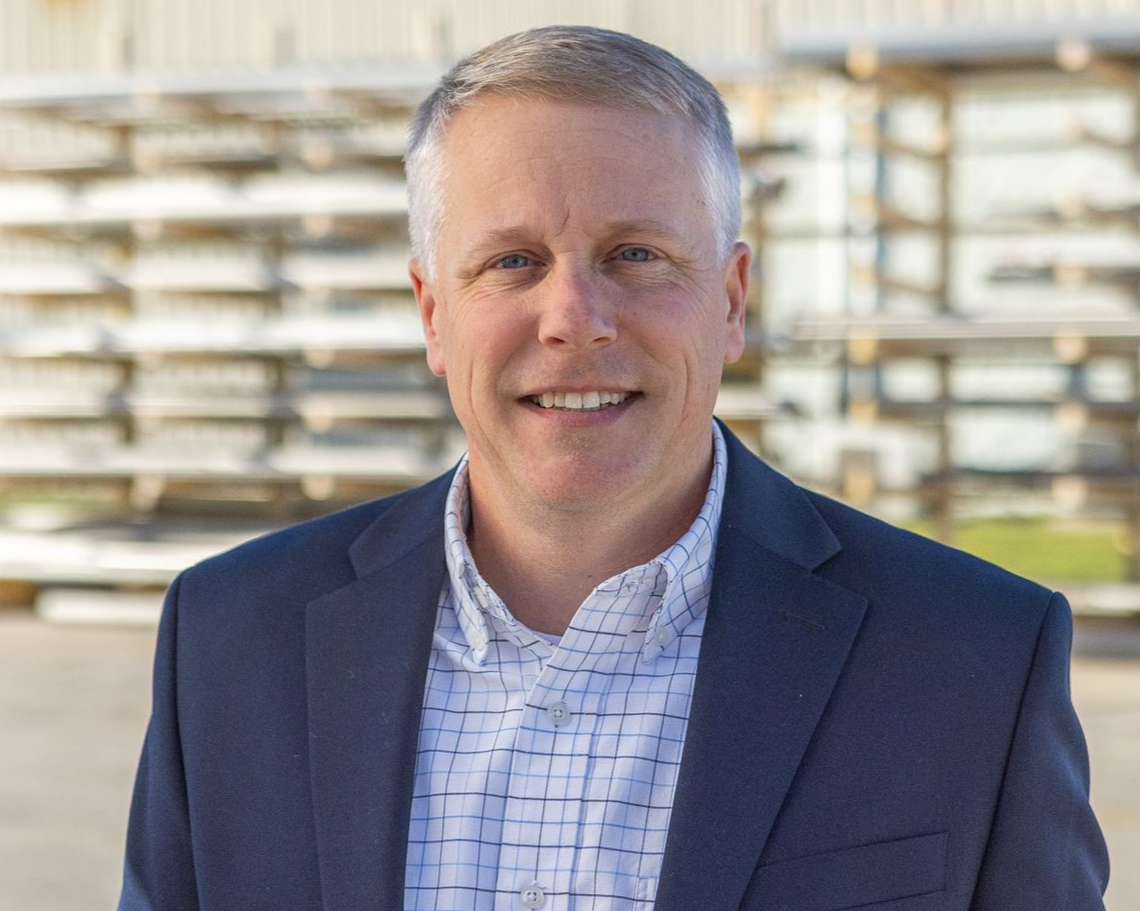 Man with short gray hair smiles, wearing a navy blazer over a white shirt, black background.