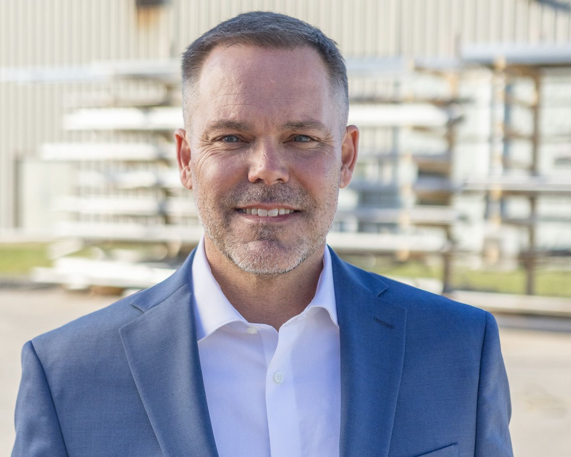 Man in dark suit, white shirt, arms crossed, looking at the viewer against a black background.