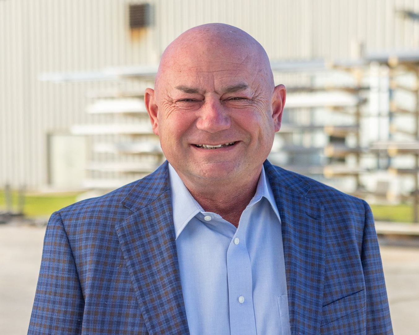 Bald man in a gray suit jacket and white shirt smiles at the camera against a mottled gray background.
