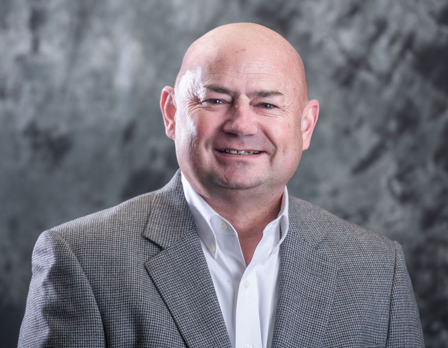 Bald man in a gray suit jacket and white shirt smiles at the camera against a mottled gray background.