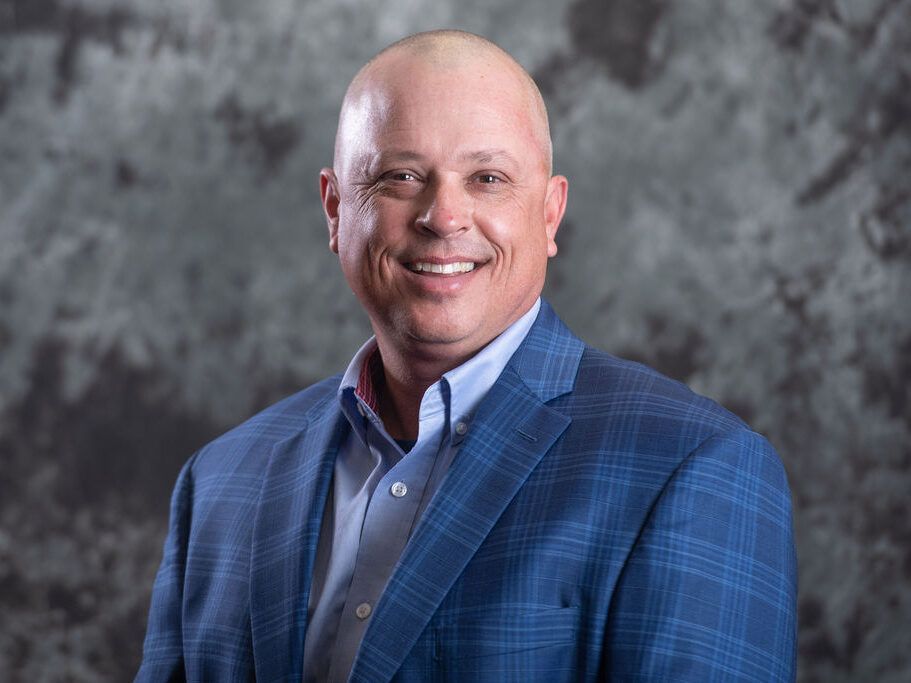 Smiling man with a shaved head in a blue plaid blazer and blue button-down shirt.