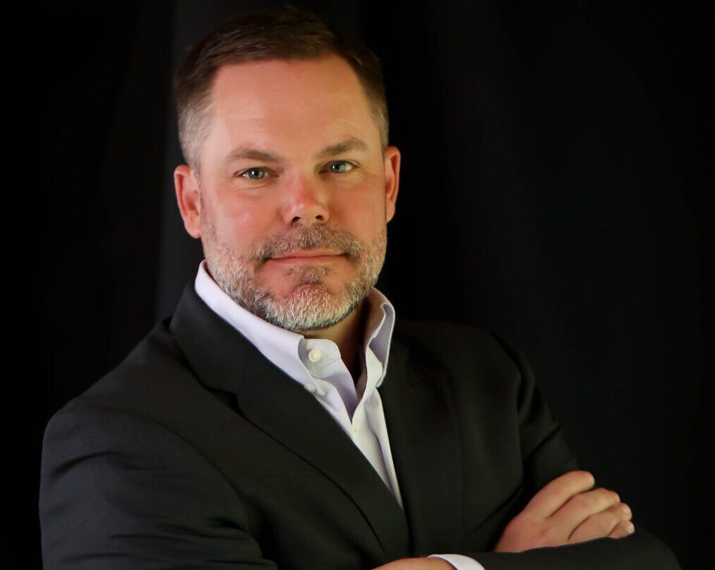 Man in dark suit, white shirt, arms crossed, looking at the viewer against a black background.