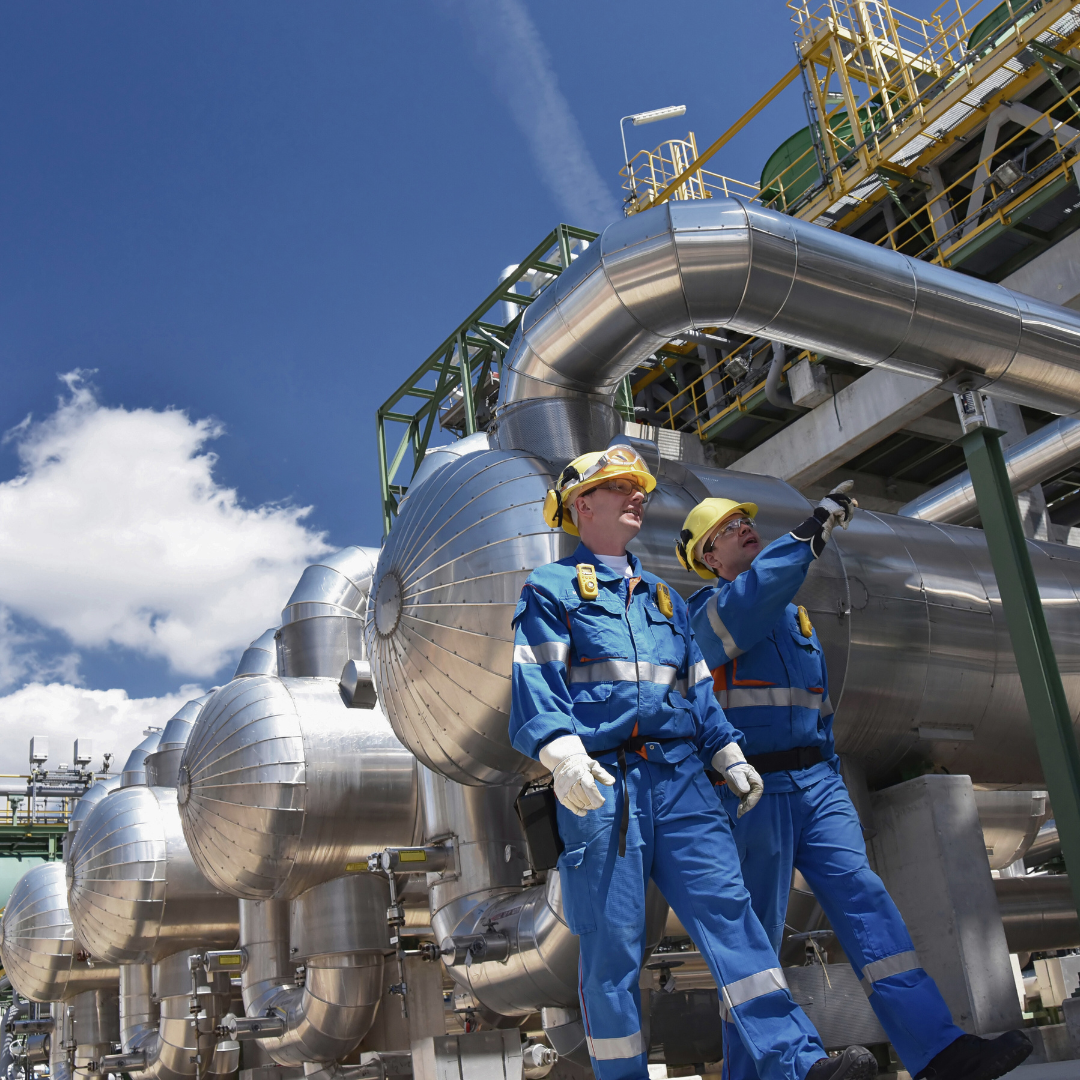 Two workers in blue coveralls and hard hats inspecting industrial equipment outdoors.