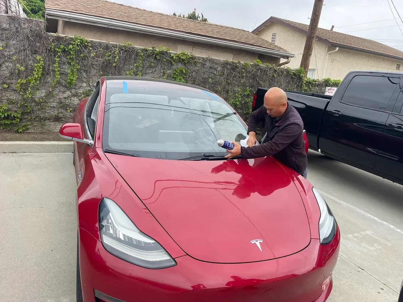 A technician applies sealant to the windshield of a red Tesla parked in an outdoor lot next to a black truck.