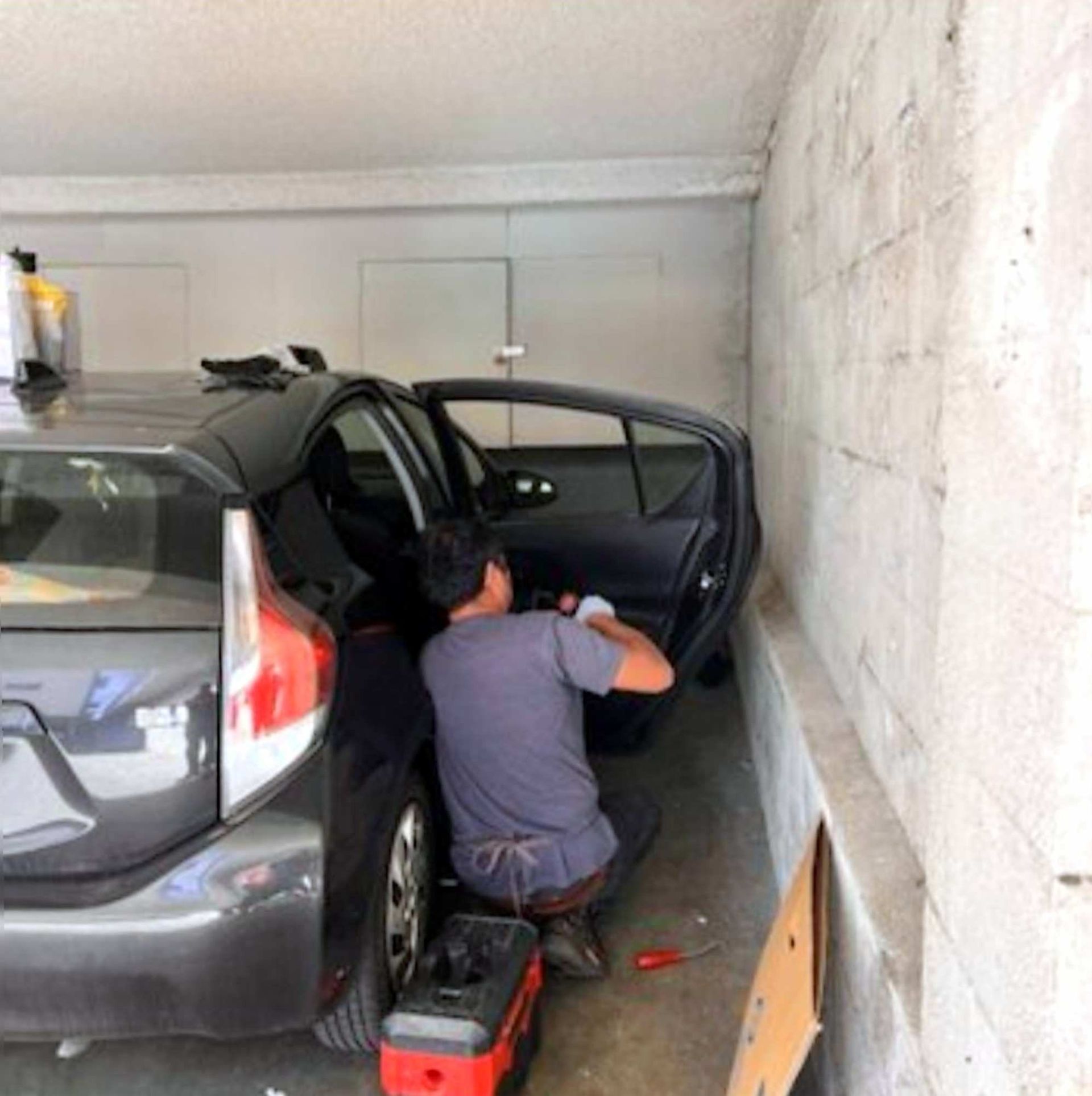 A person wearing a grey shirt works on the inside of a grey car door while parked in a concrete garage.