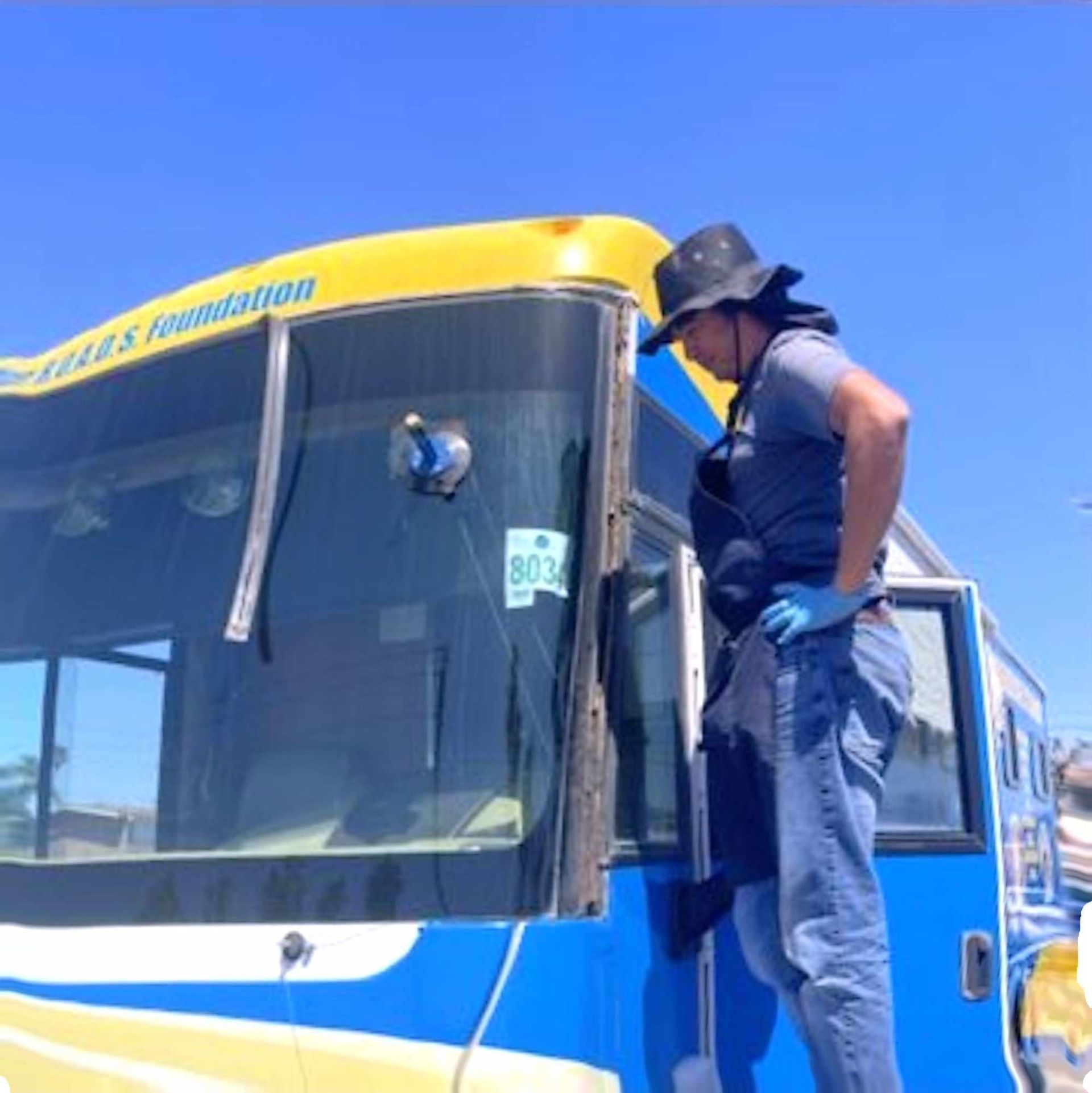 A person in a hat and blue gloves stands on the side of a blue and yellow bus while working on the windshield.