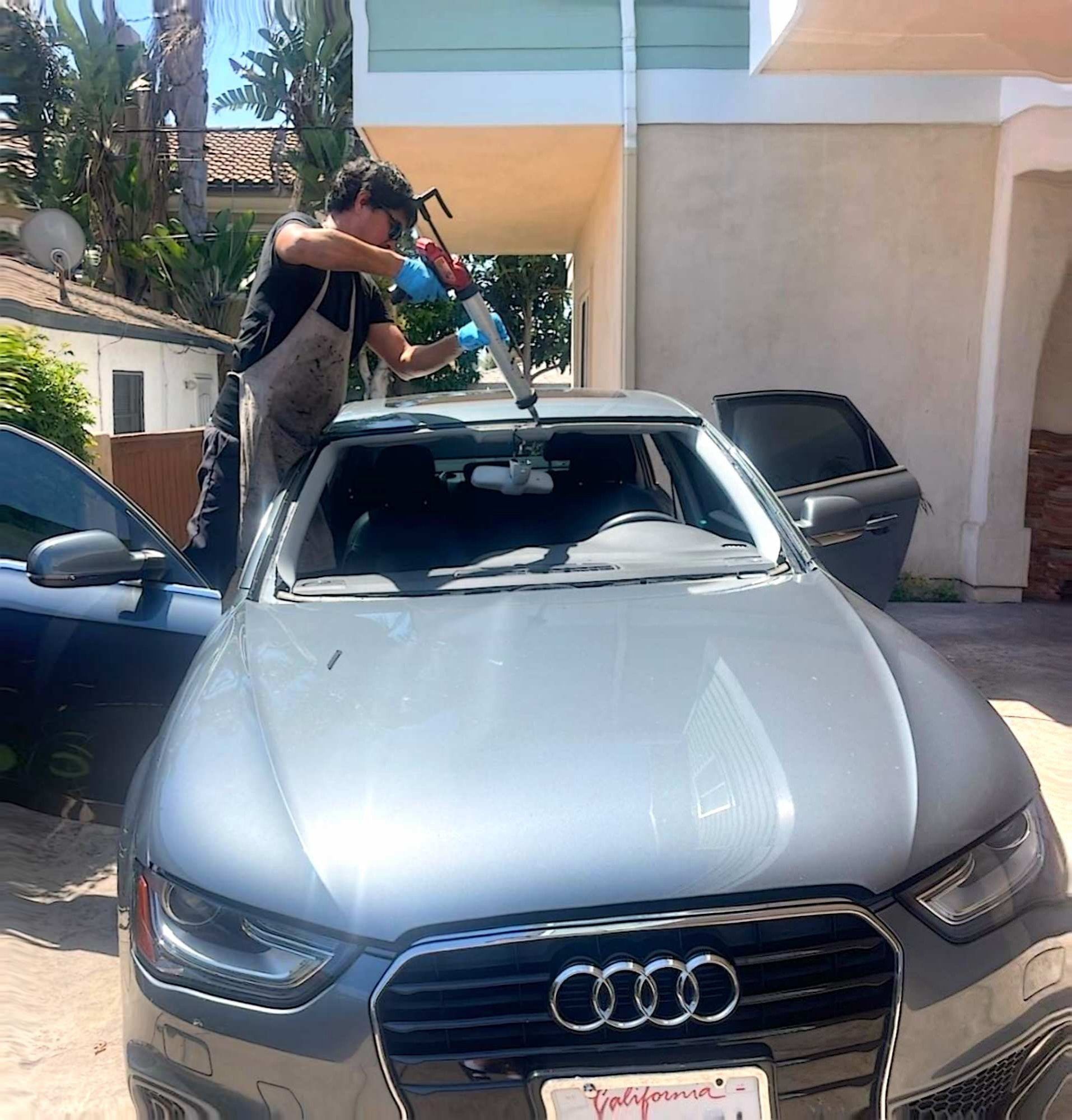 A person wearing a tan apron and blue gloves uses a tool to install a new windshield on a gray Audi car outdoors.
