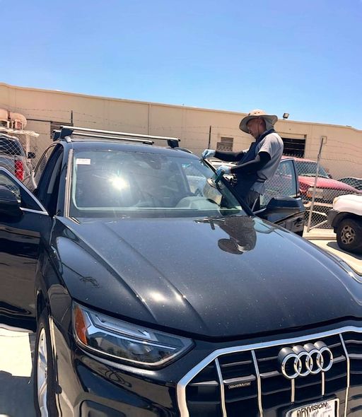 A person wearing a wide-brimmed hat works on the windshield of a black Audi parked at a facility.