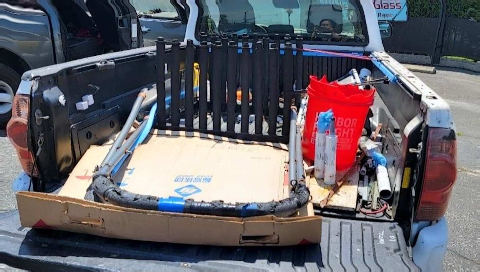 A black rod rack stands in the open bed of a white pickup truck, alongside a red bucket, cardboard, and metal pipes.