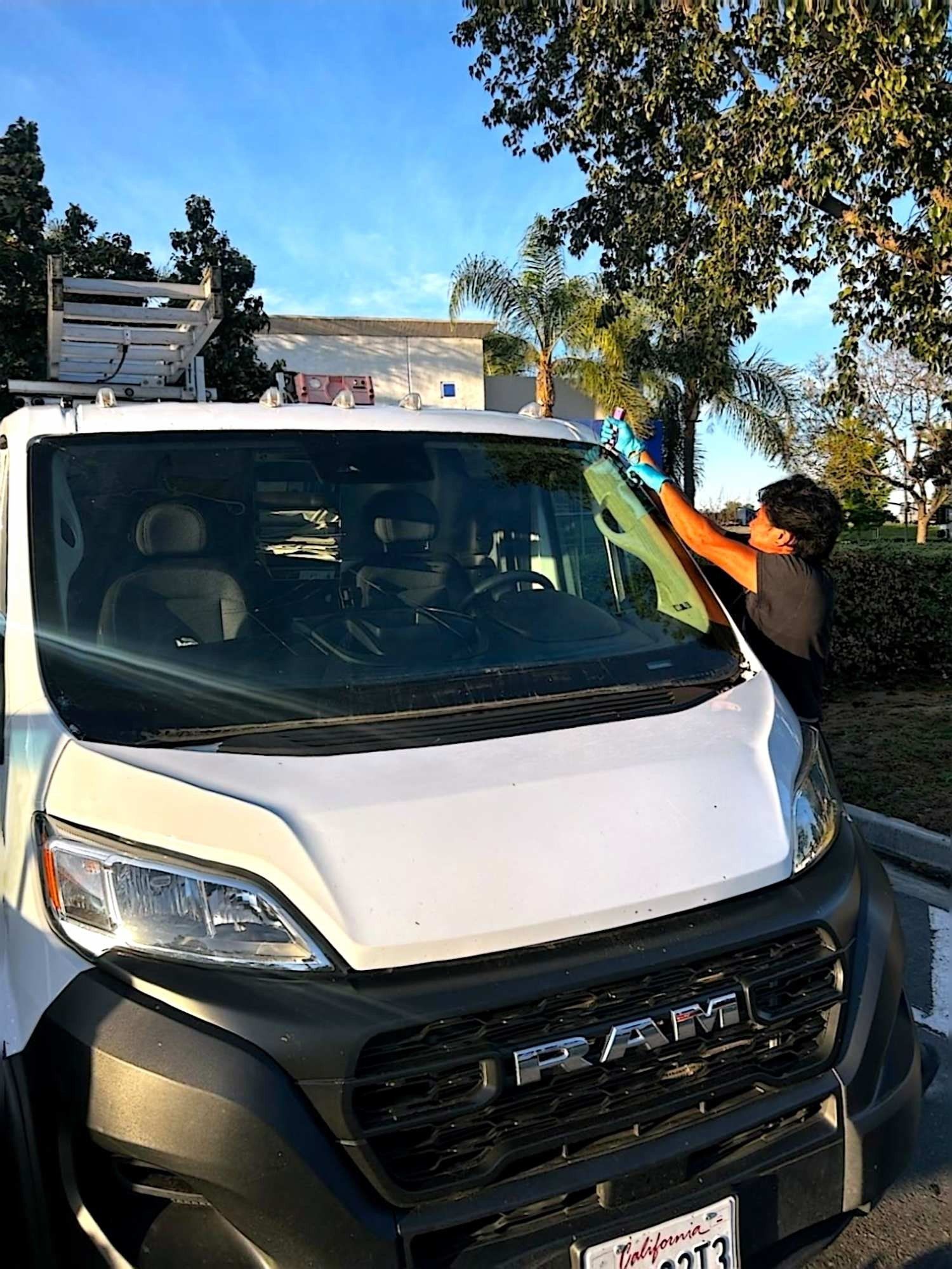 A worker in blue gloves wipes the windshield of a white Ram cargo van parked outdoors under a sunny sky.