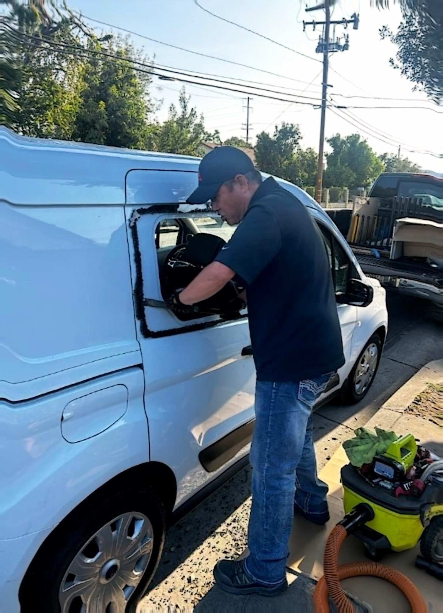 A person in a dark shirt and jeans works on the back window of a white van, using a shop vacuum on a sunny street.