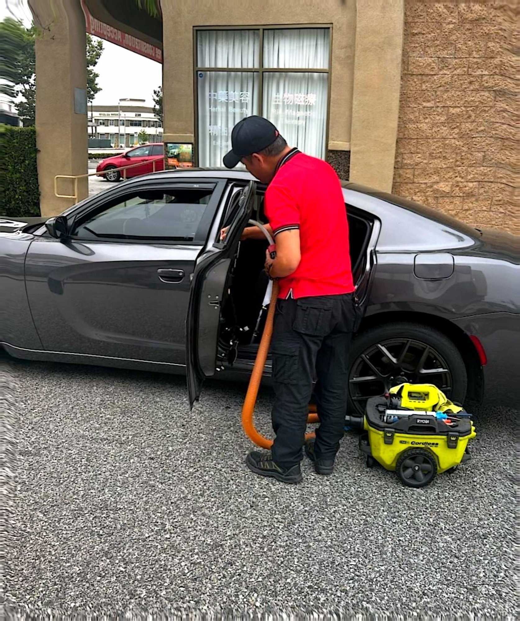 A person in a red shirt vacuums the interior of a gray sedan parked on a gravel surface.
