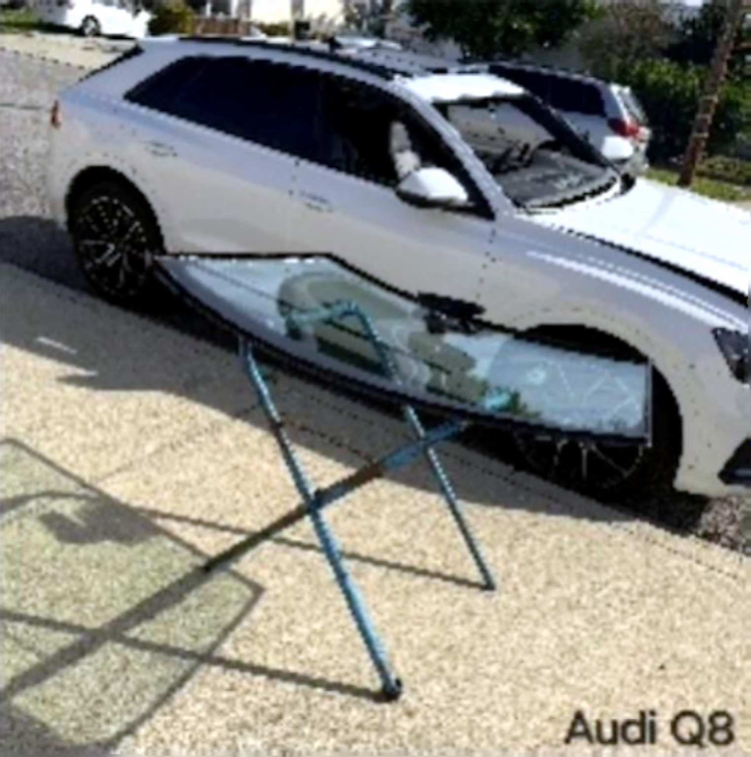 A new windshield sits on a stand in front of a white Audi Q8 parked on a paved driveway.