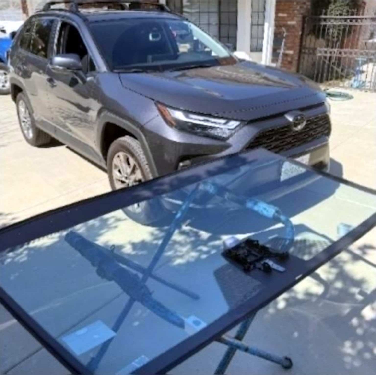 A new replacement windshield rests on a stand in front of a dark grey Toyota RAV4 parked in a driveway.