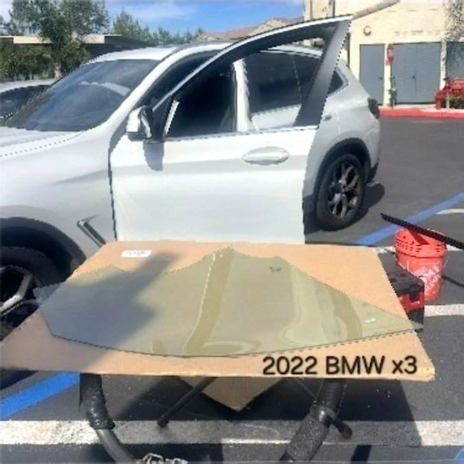 A replacement car window pane sits on a cardboard box in front of a white 2022 BMW X3 with its rear door window missing.
