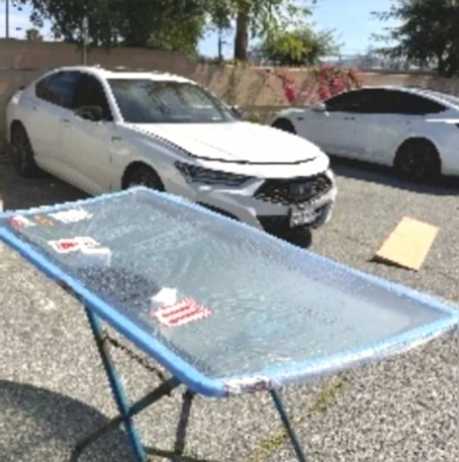 A replacement car windshield sits on a portable stand in a parking lot, with white cars parked in the background.