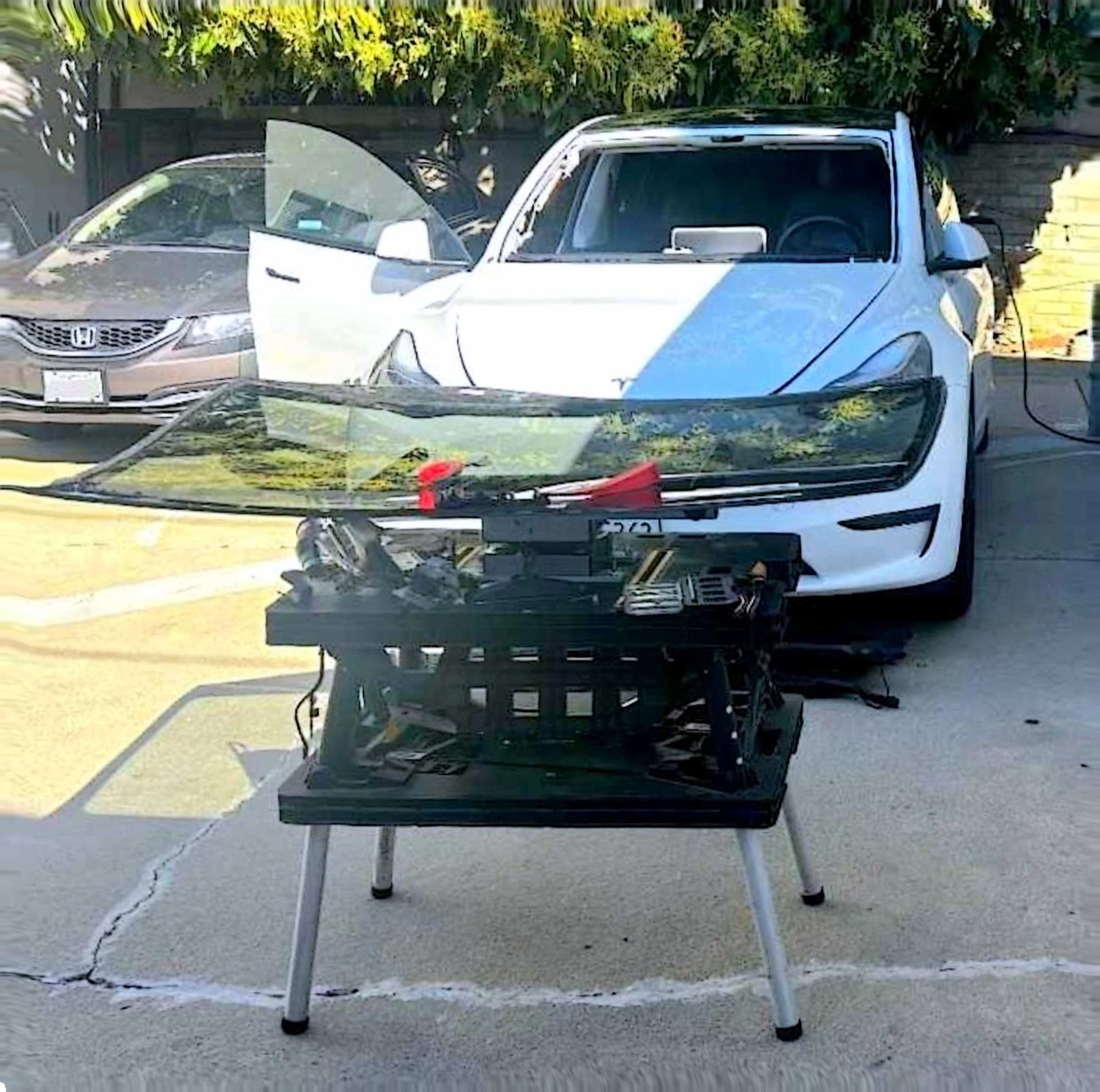 A replacement windshield sits on a portable stand in front of a white car parked in a driveway.