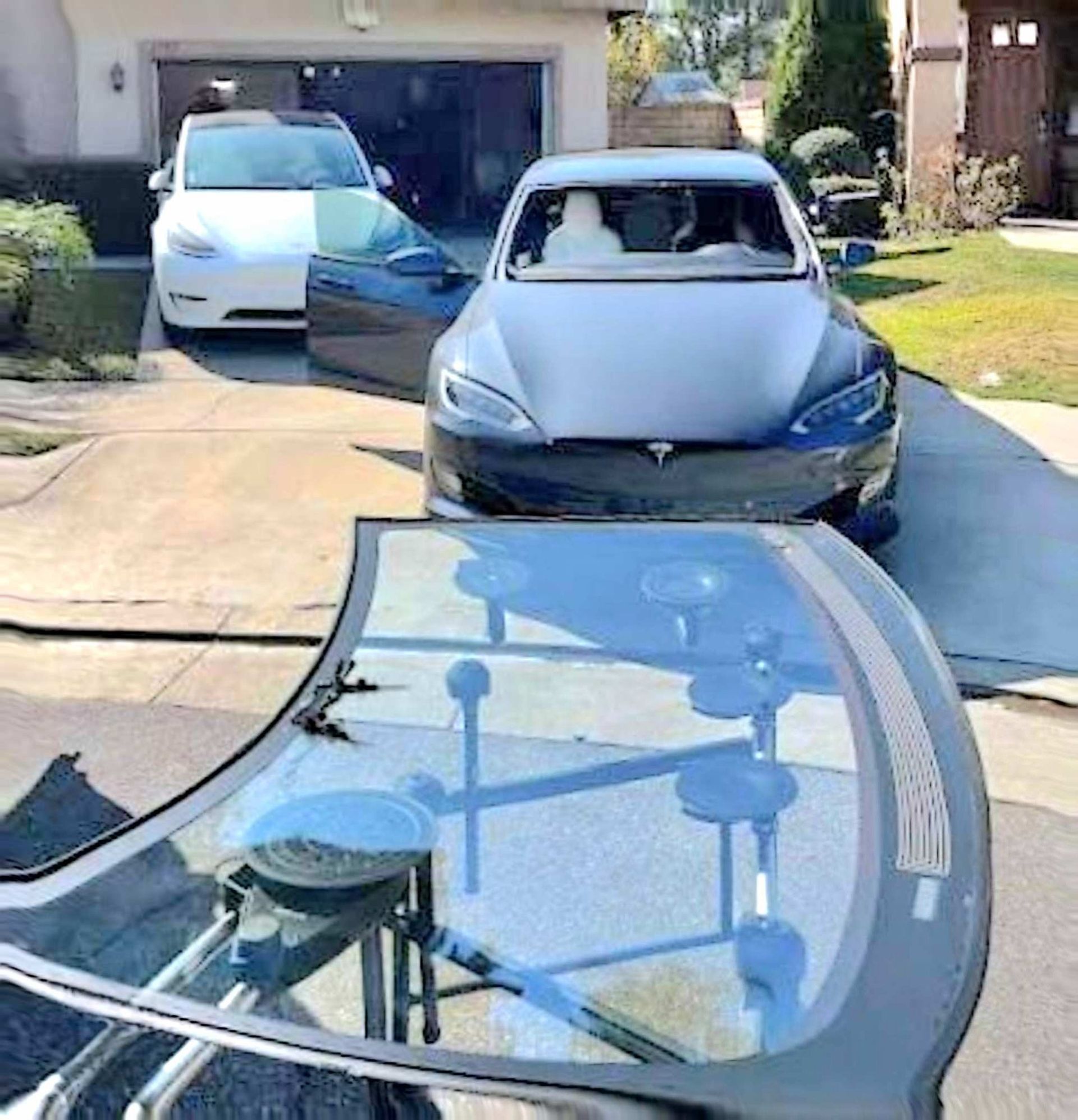 A Tesla windshield sits on a suction cup stand in the foreground, with a grey Tesla and a white Tesla parked behind it.