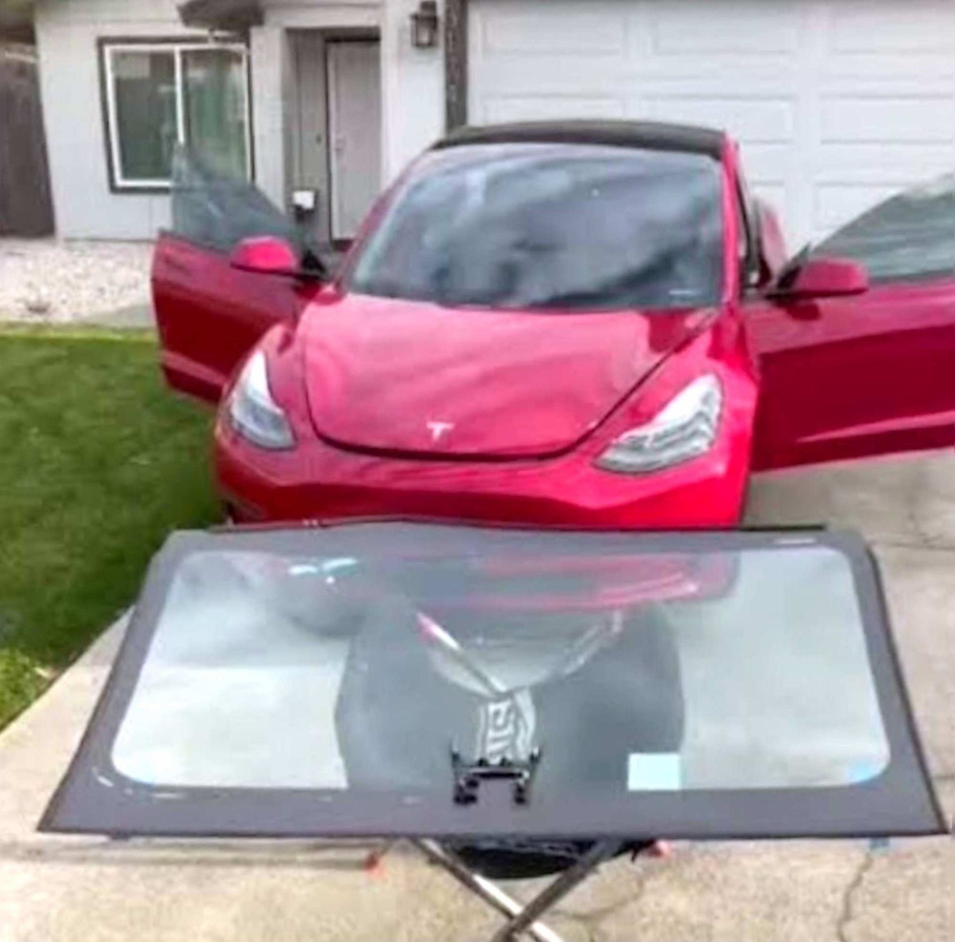 A red Tesla with doors open parked in a driveway in front of a new glass windshield on a stand.