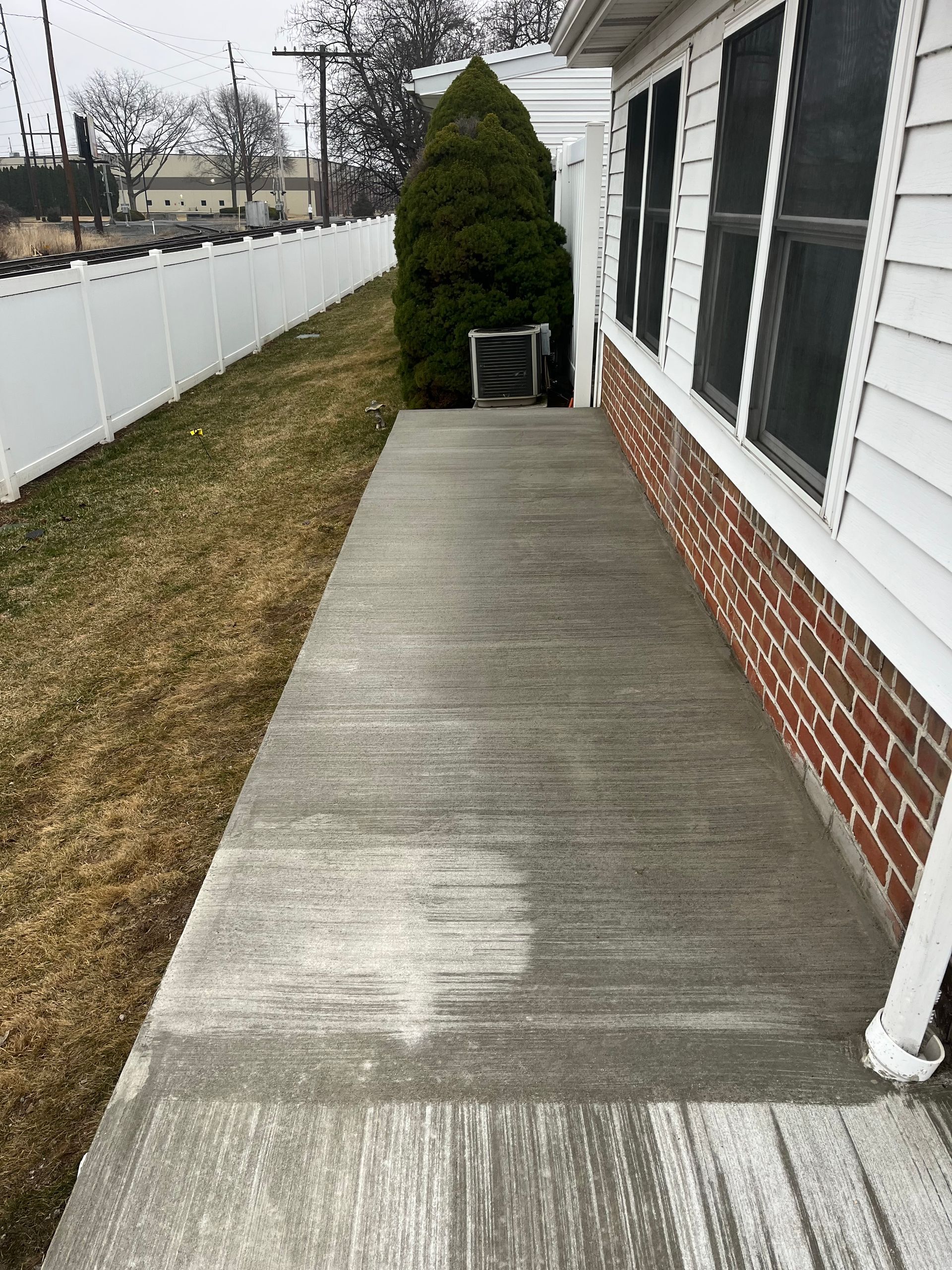 A long, gray concrete walkway next to a white house, surrounded by grass and a white fence.