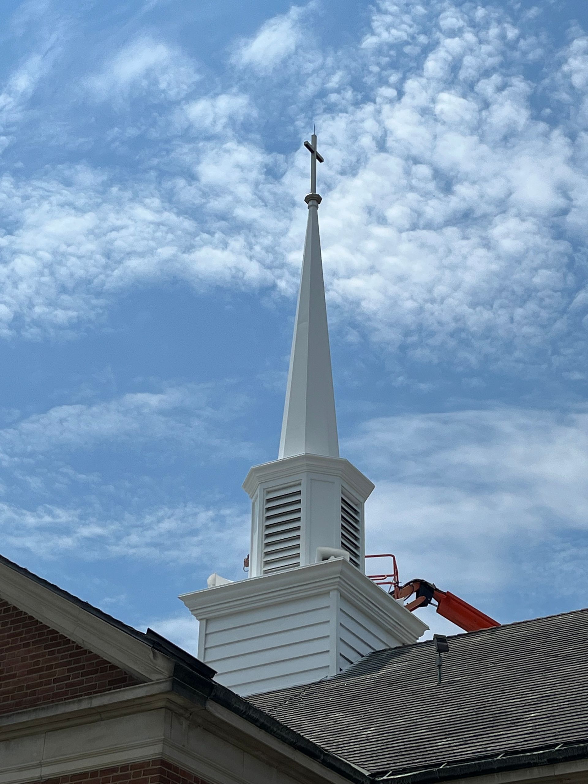 White church steeple with cross against a blue sky; worker in orange lift is visible on the roof.