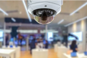 Security camera dome inside a blurred retail store, white tables, customers browsing.