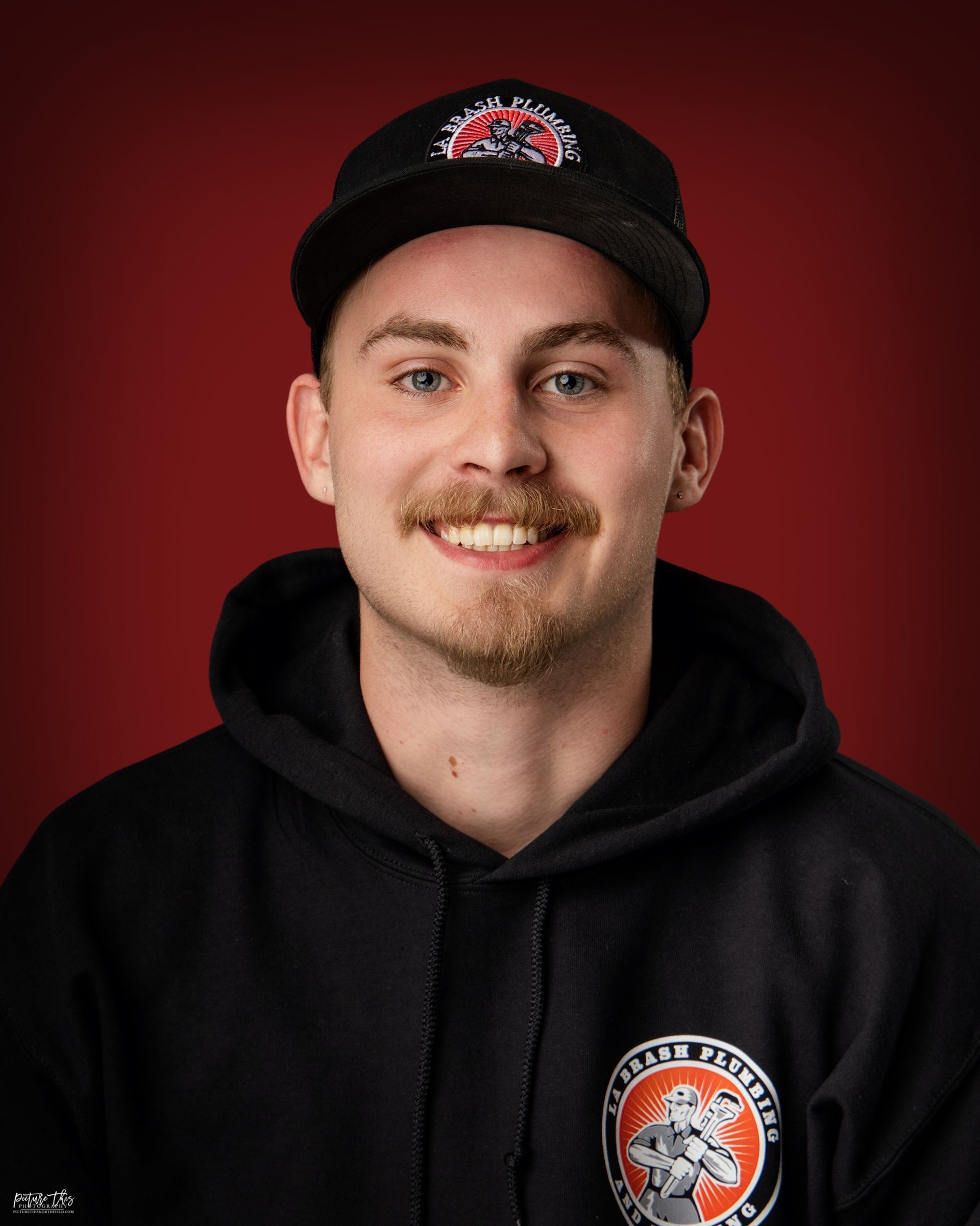 Young man smiling, wearing a black hoodie and baseball cap, standing against a red backdrop.