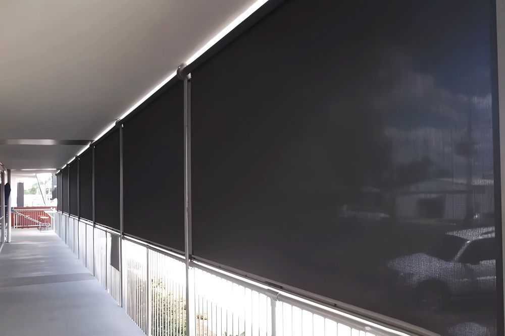 Dark roller shades line a covered outdoor walkway, blocking sunlight.