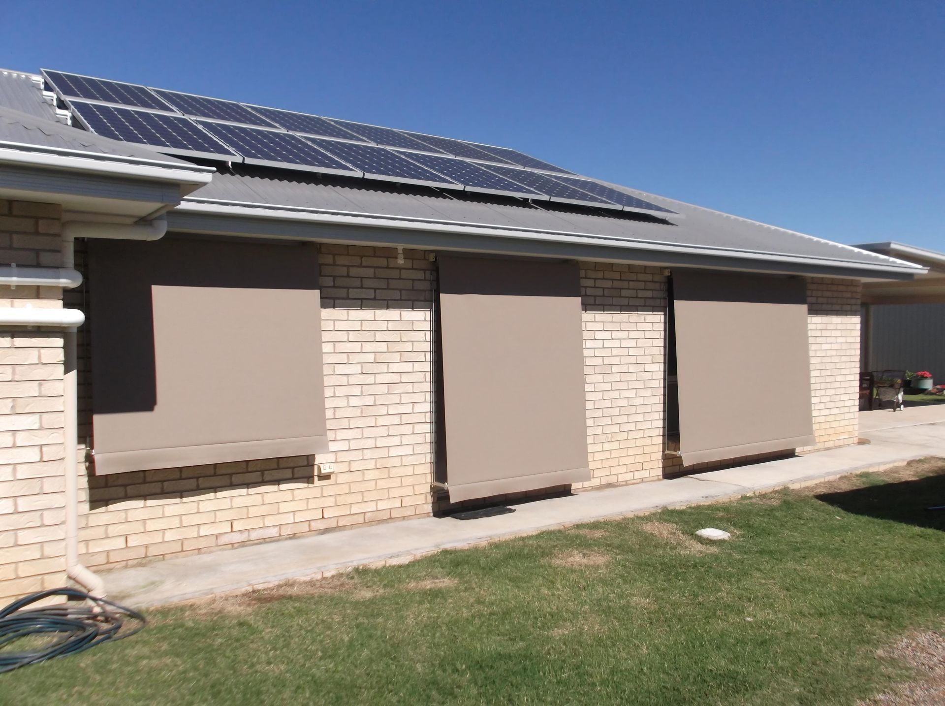 Three beige rectangular sun shades on a brick wall of a house with solar panels on the roof.
