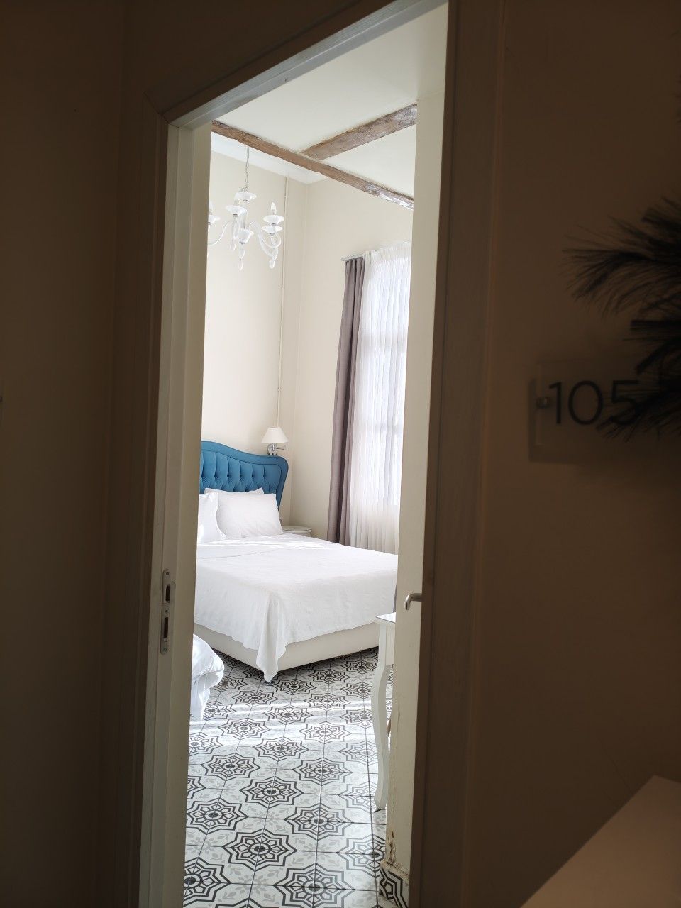 Μια πόρτα υπνοδωματίου με την ένδειξη 105, που δείχνει ένα λευκό κρεβάτι με γαλαζοπράσινο κεφαλάρι και δάπεδο με πλακάκια με σχέδια.