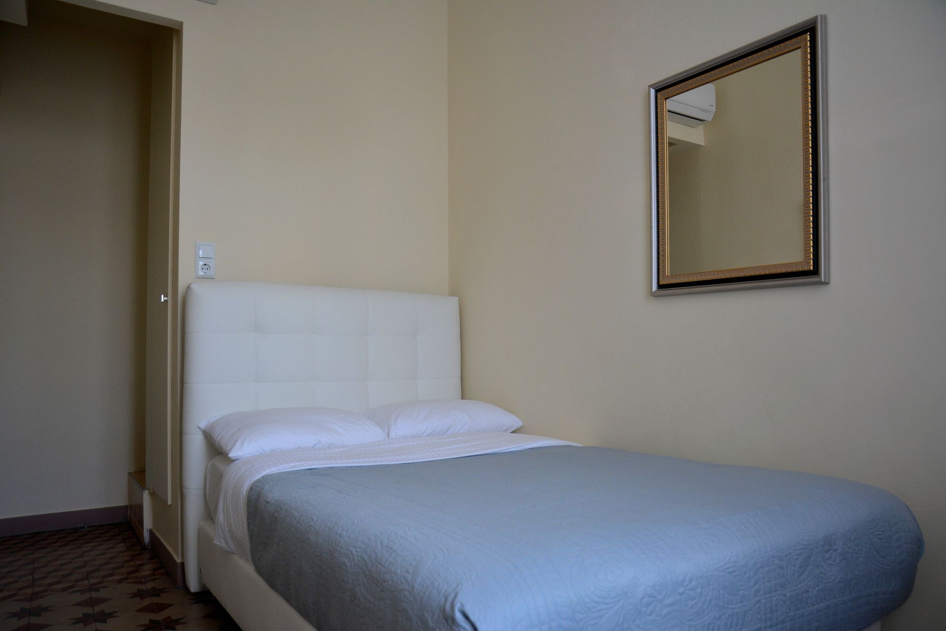 Ένα υπνοδωμάτιο με λευκό καπιτονέ κεφαλάρι κρεβατιού, γκρι κλινοσκεπάσματα και έναν τετράγωνο καθρέφτη στον τοίχο.