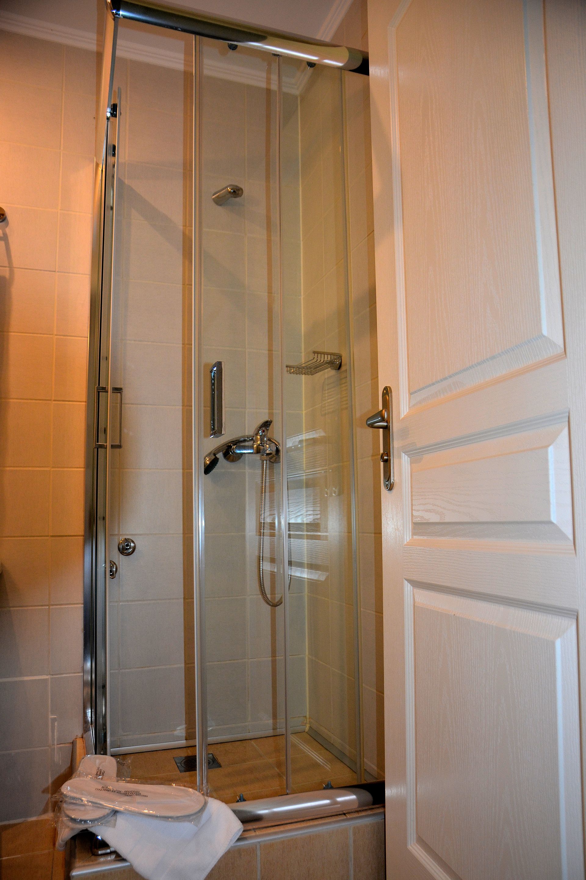 Μια καμπίνα ντους με γυάλινες πόρτες, ασημένια κεφαλή ντους και μια βρύση στο εσωτερικό, που βρίσκεται δίπλα σε μια λευκή πόρτα σε ένα μπάνιο.