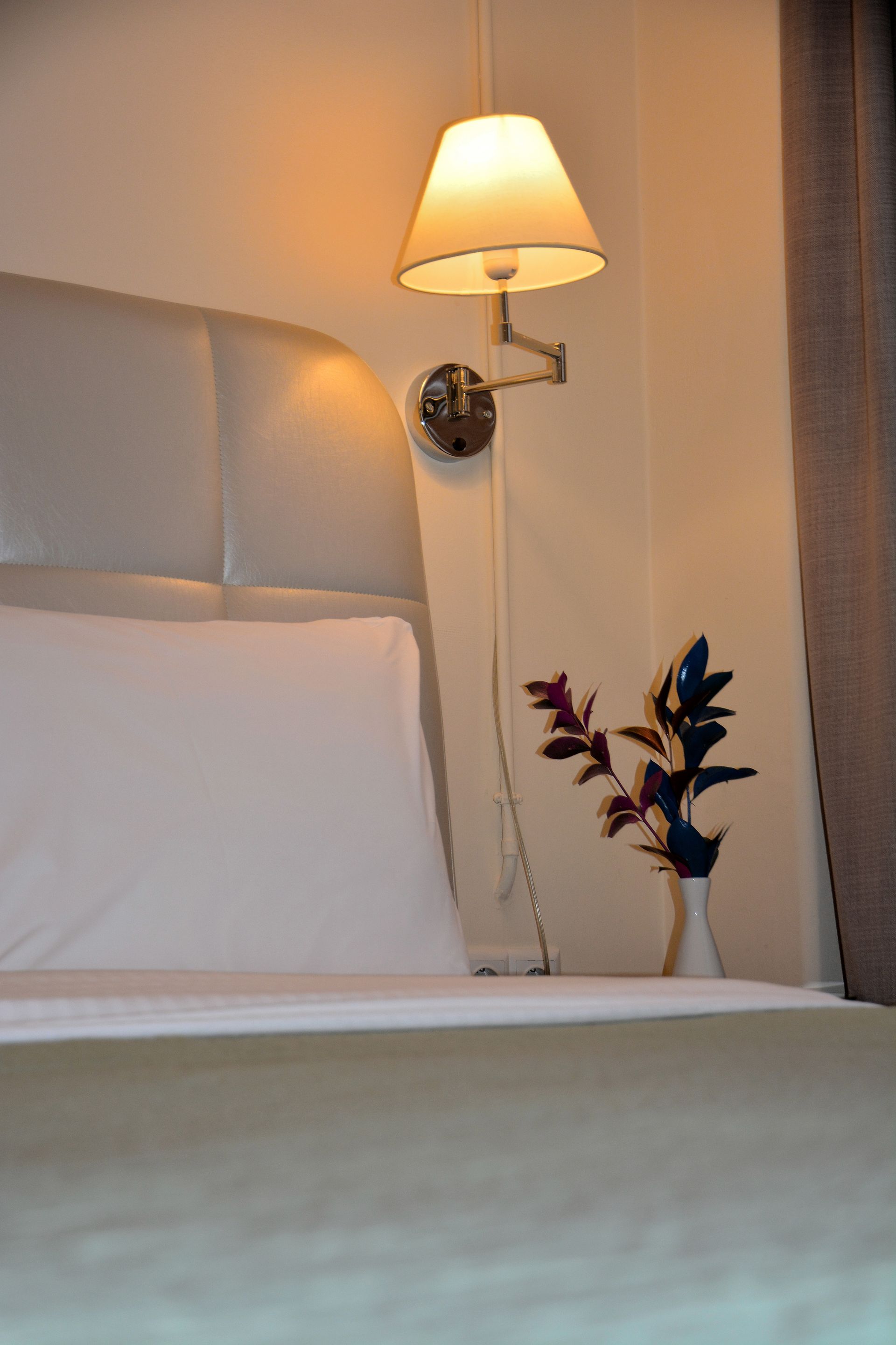 Μια αμυδρά φωτισμένη γωνιά δωματίου ξενοδοχείου με ένα γκρι κεφαλάρι με επένδυση, μια επιτοίχια λάμπα και ένα βάζο με σκούρα φυλλώματα.