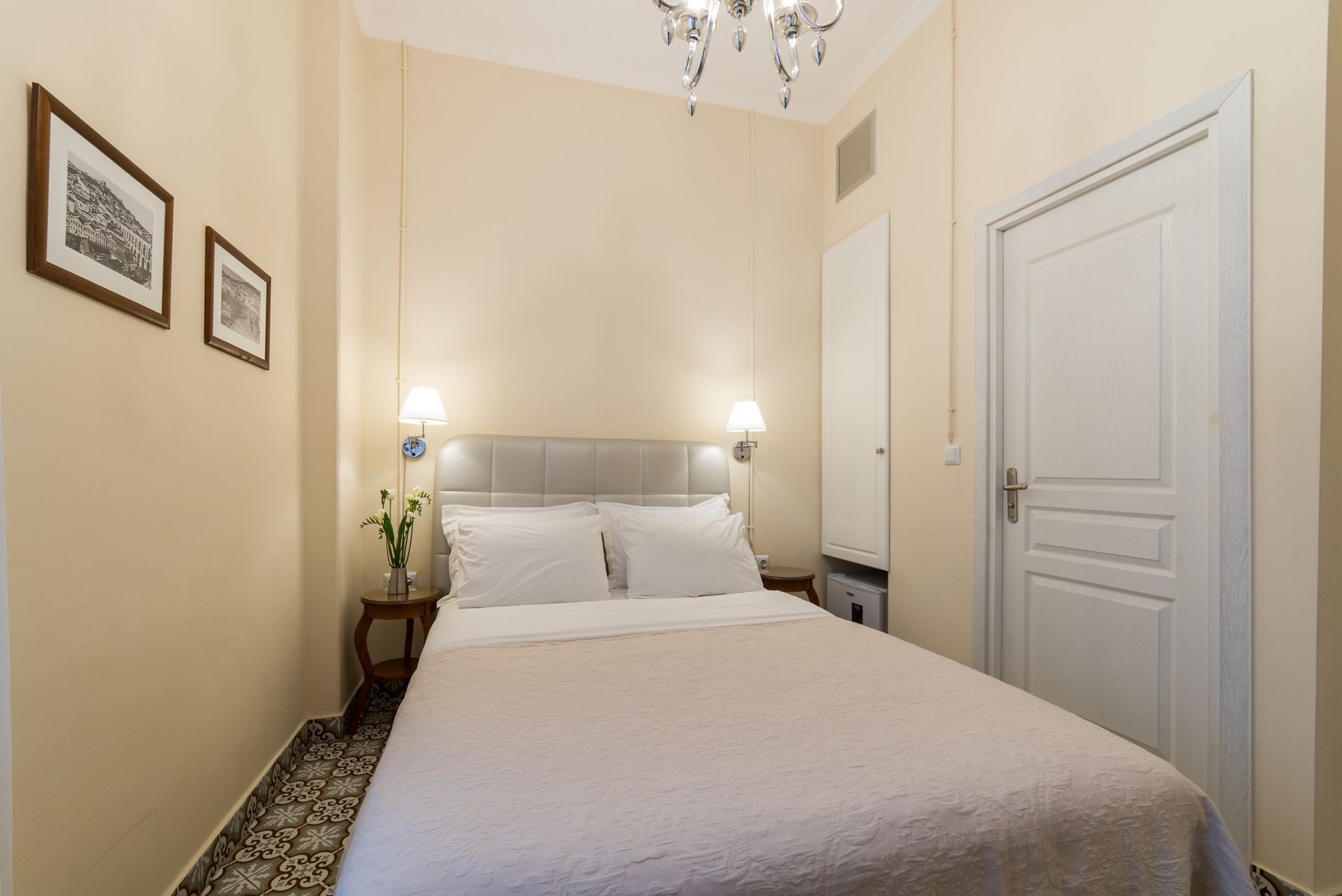 Ένα μικρό, προσεγμένο υπνοδωμάτιο με διπλό κρεβάτι, ανοιχτόχρωμους μπεζ τοίχους, λευκές πόρτες και έναν κρυστάλλινο πολυέλαιο.