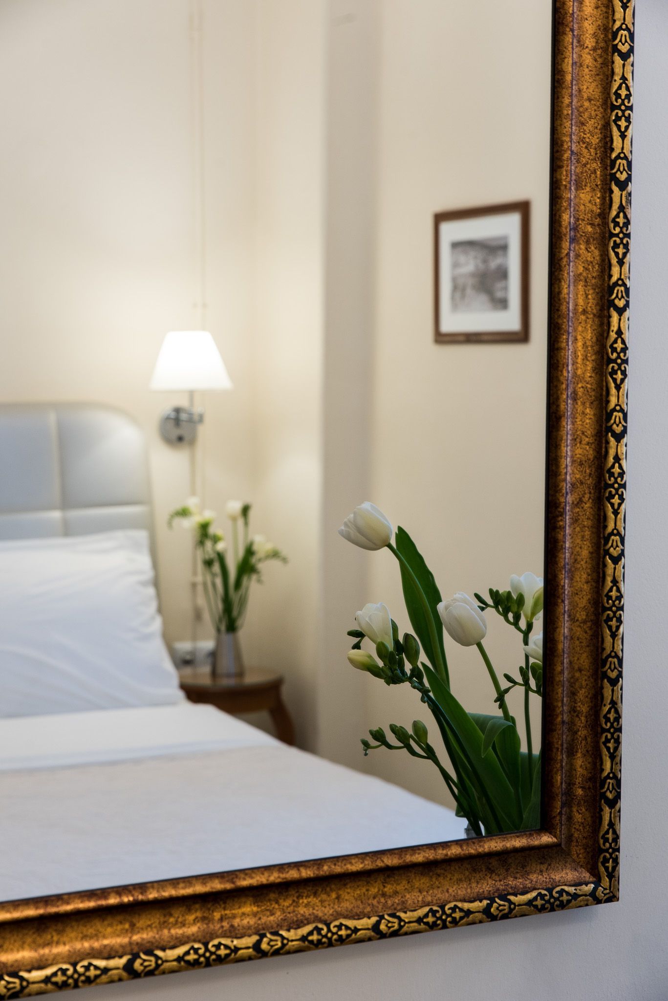 Ένας περίτεχνος καθρέφτης με χρυσό σκελετό που αντανακλά ένα τμήμα ενός υπνοδωματίου με ένα λευκό κρεβάτι, ένα μικρό βάζο με λευκά λουλούδια και ένα φωτιστικό.