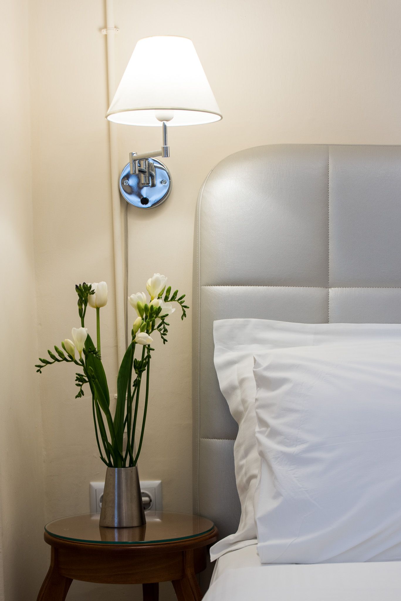 Ένα λευκό επιτοίχιο φωτιστικό κρέμεται πάνω από ένα μικρό ξύλινο βοηθητικό τραπεζάκι που κρατάει ένα βάζο με λευκά λουλούδια δίπλα σε ένα γκρι κεφαλάρι κρεβατιού.