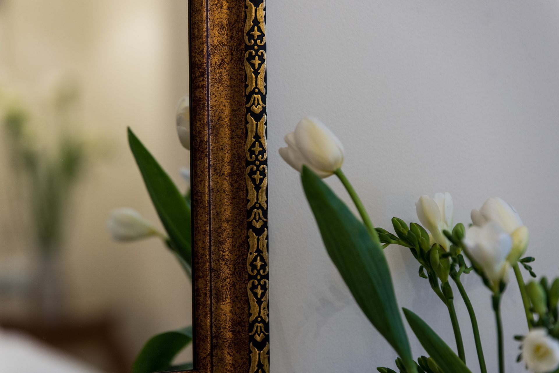 Λευκά λουλούδια τοποθετημένα δίπλα σε έναν περίτεχνο, χρυσόσκελετο καθρέφτη, που αντανακλούν τους μίσχους και το πράσινο σε ένα απαλό, φωτεινό δωμάτιο.