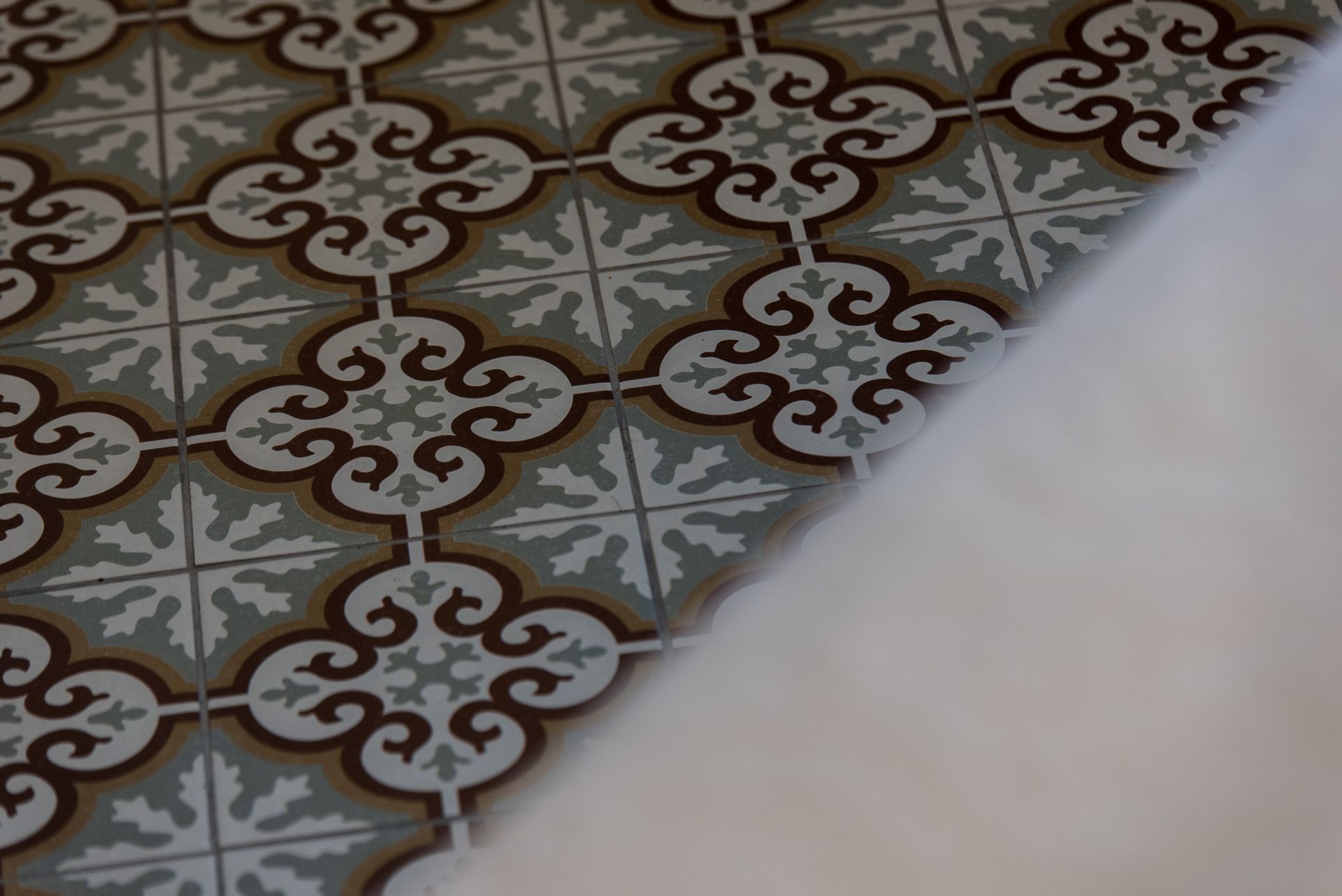 Ένα πλακόστρωτο δάπεδο με ένα επαναλαμβανόμενο καφέ και κρεμ γεωμετρικό, λουλουδάτο μοτίβο δίπλα σε μια θολή λευκή υφασμάτινη επιφάνεια.
