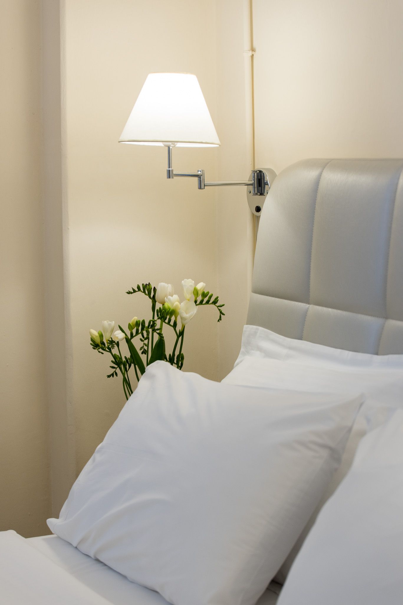 Ένα λευκό φωτιστικό σε αρθρωτό μπράτσο κρέμεται πάνω από ένα γκρι επενδεδυμένο κεφαλάρι κρεβατιού με ένα μικρό μπουκέτο λουλούδια δίπλα του.