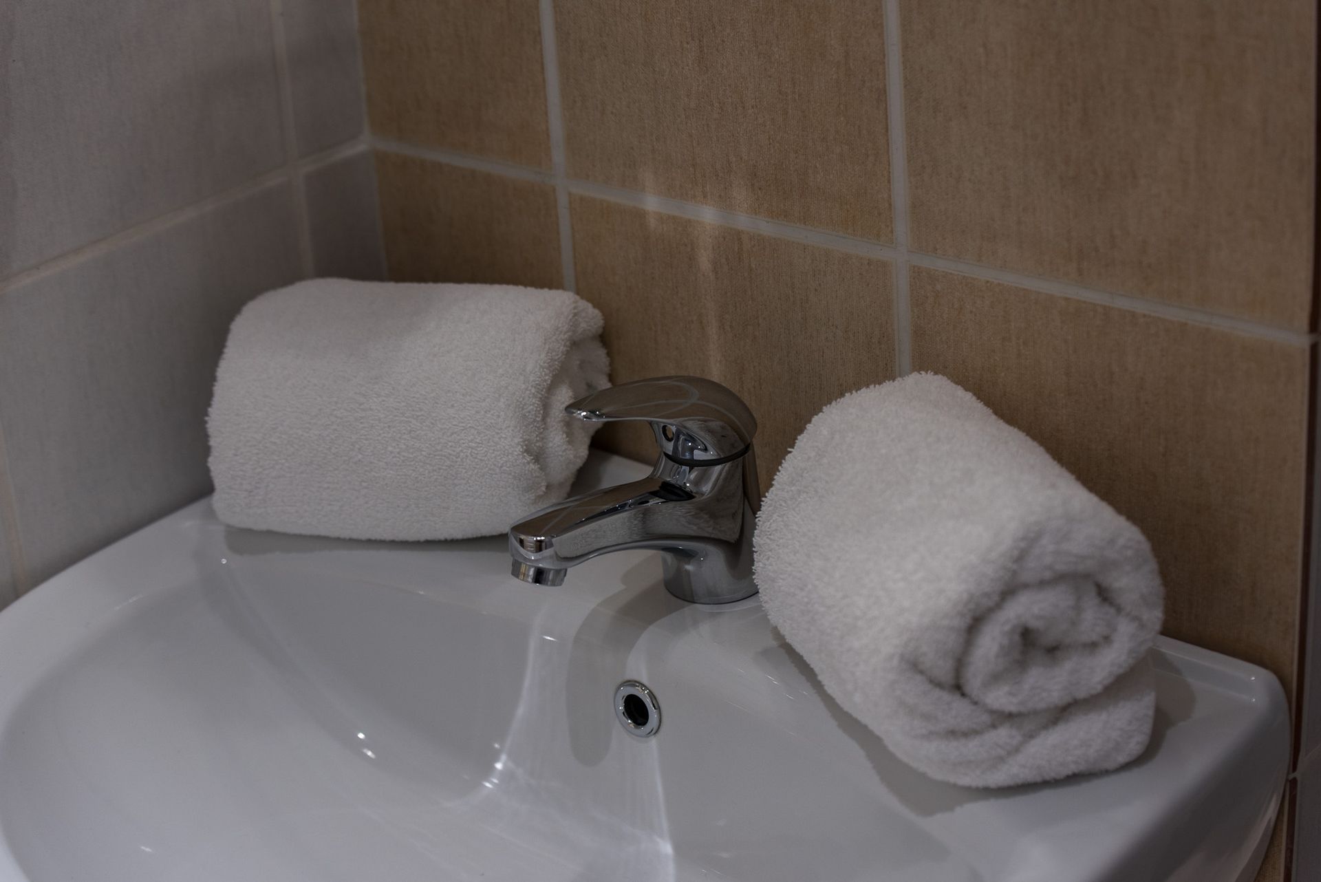 Δύο διπλωμένες λευκές πετσέτες ακουμπούν σε έναν λευκό νιπτήρα εκατέρωθεν μιας μεταλλικής βρύσης σε τοίχους με καφέ πλακάκια.