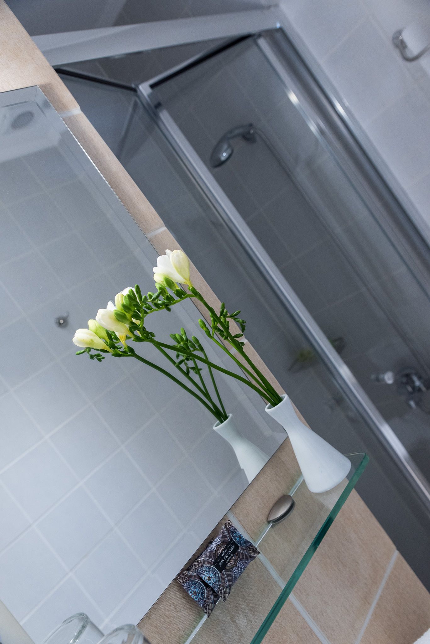 Ένα βάζο με λευκά λουλούδια κάθεται σε ένα γυάλινο ράφι μπροστά από έναν καθρέφτη του μπάνιου, αντανακλώντας μια καμπίνα ντους.