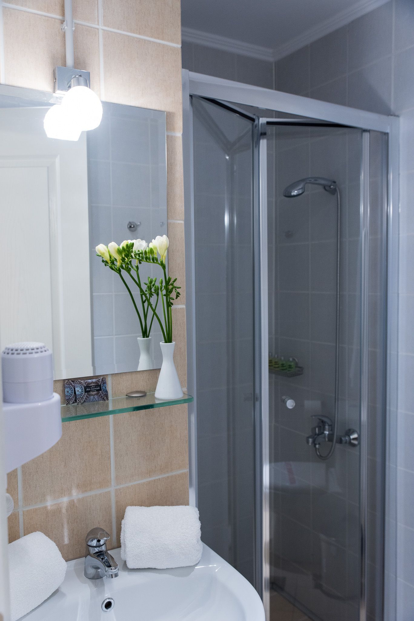 Ένα μπάνιο με λευκό νιπτήρα, πλακάκια στους τοίχους, έναν καθρέφτη με λουλούδια και μια γυάλινη καμπίνα ντους.