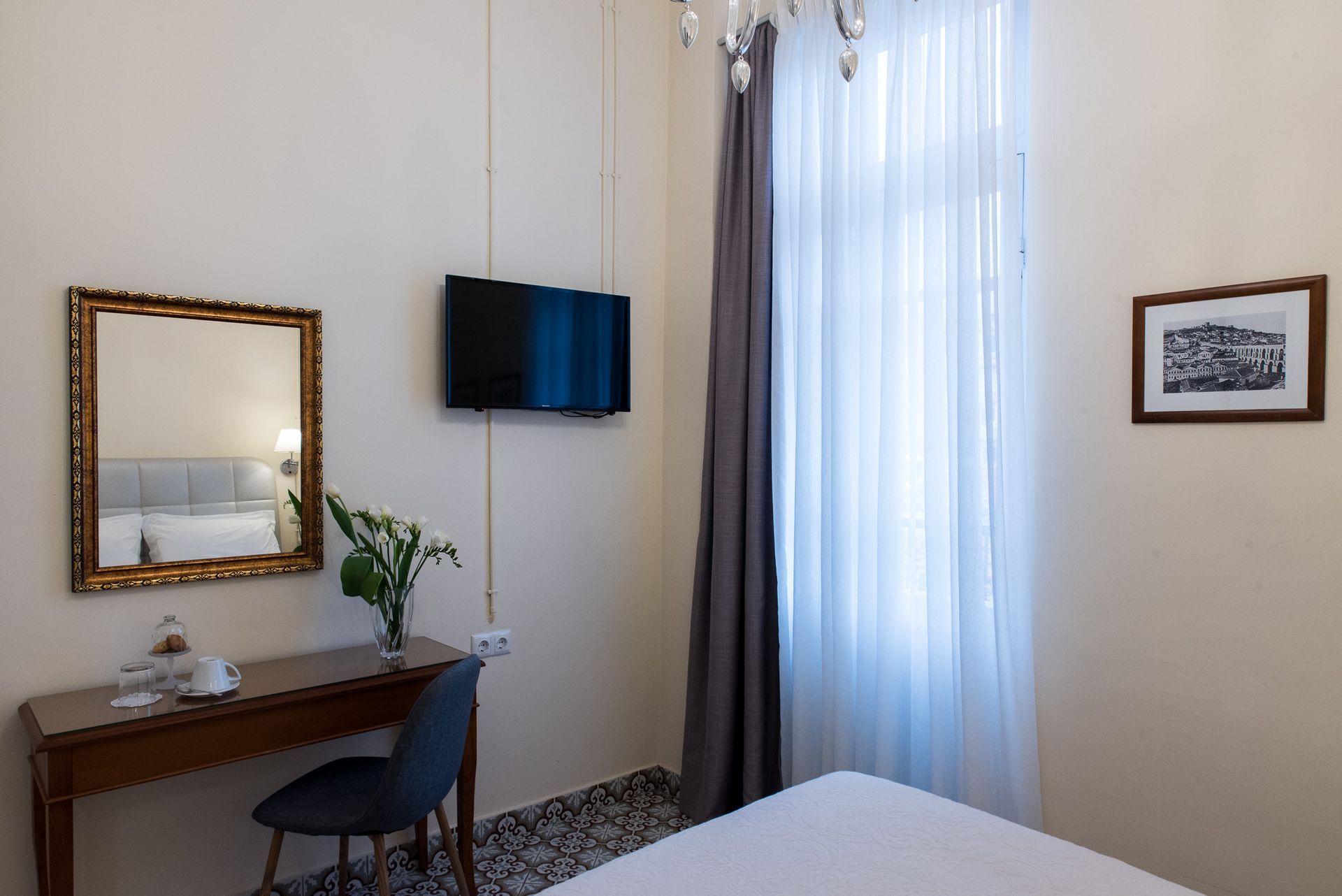 Ένα δωμάτιο ξενοδοχείου με γραφείο, καθρέφτη, επιτοίχια τηλεόραση, παράθυρο με διαφανείς κουρτίνες και μια πλαισιωμένη εκτύπωση στον τοίχο.